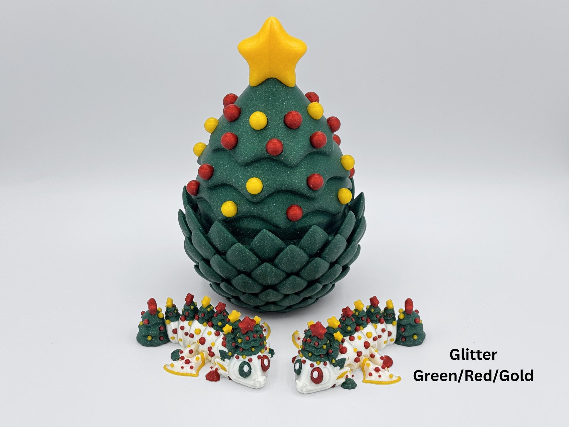 A glitter green 3D printed Christmas tree egg with glitter red and yellow ornaments and a yellow star topper, displayed with two white articulated Christmas tree dragons decorated in matching green, red, and gold tree details on a white background for the Christmas dragon and egg holiday tabletop decor listing.