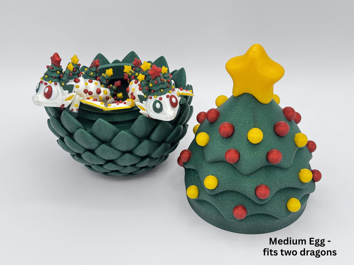 A medium 3D printed Christmas egg in glitter green with red and yellow ornaments, shown open with two white articulated Christmas tree dragons posed inside, displayed next to the matching closed tree lid with a yellow star topper on a white background for the Christmas dragon holiday tabletop decor listing.